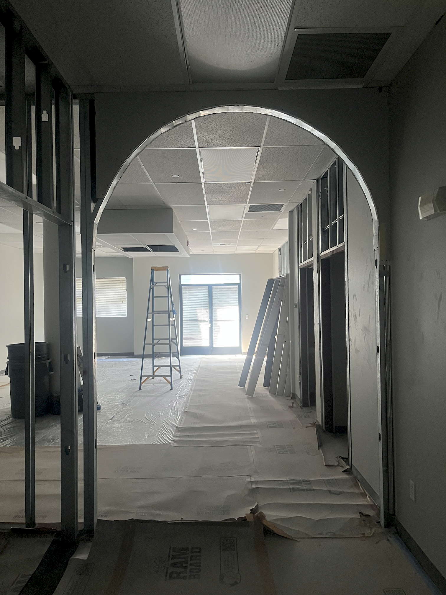 SDG pre-construction 3 A view into an office space undergoing remodeling. Directly forward is a tall arch, empty doorways to the right along with the doors to be hung in them, an open ladder, and the back doors. To the left is the open metal framework of a wall in progress, with a couple buckets visible just part that.