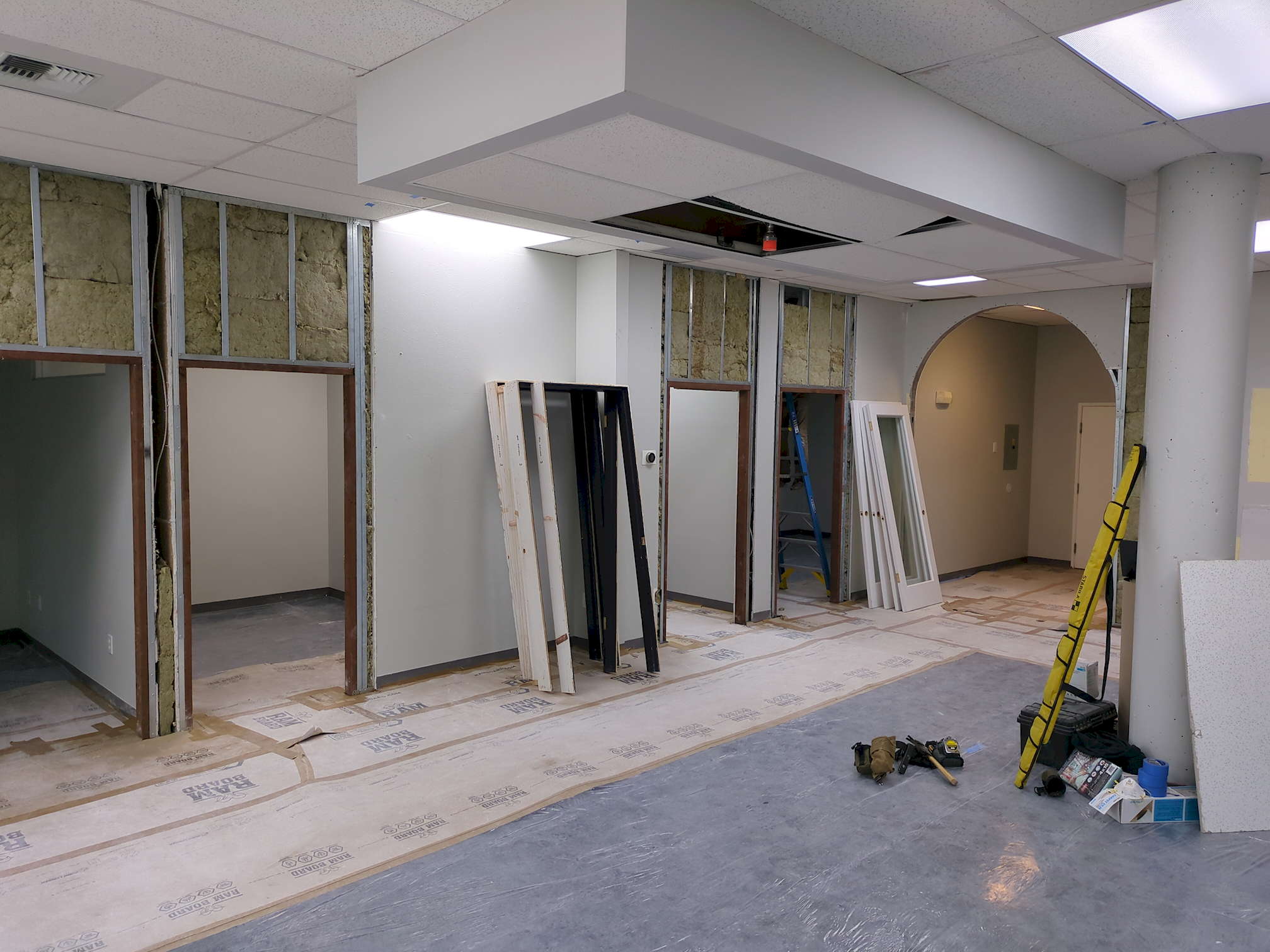 SDG pre-construction 4 A later view of the same office from the other side. One acoustic ceiling tile is ajar. Another is missing, revealing a newly installed sprinkler head. The door frames have been partially replaced, revealing metal framework stuffed with insulation. The floor is completely covered cardboard and clear plastic, and some building materials lean against a column toward the right.
